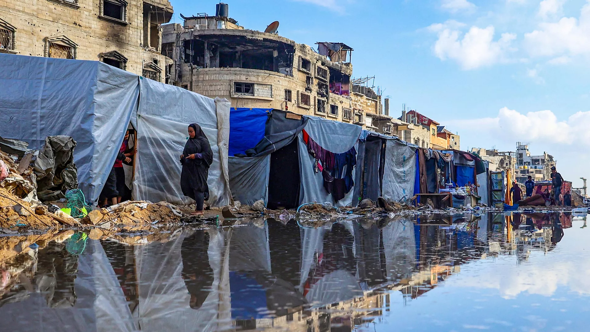 Người Palestine đi quanh vũng nước trước những tòa nhà và lều trại bị phá hủy ở Khan Yunis, Palestine