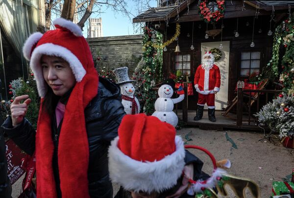 Một phụ nữ và em bé đội mũ lễ hội đi ngang qua nhân viên mặc đồ ông già Noel đang đón khách tại chợ Giáng sinh theo phong cách Pháp ở Bắc Kinh, Trung Quốc - Sputnik Việt Nam