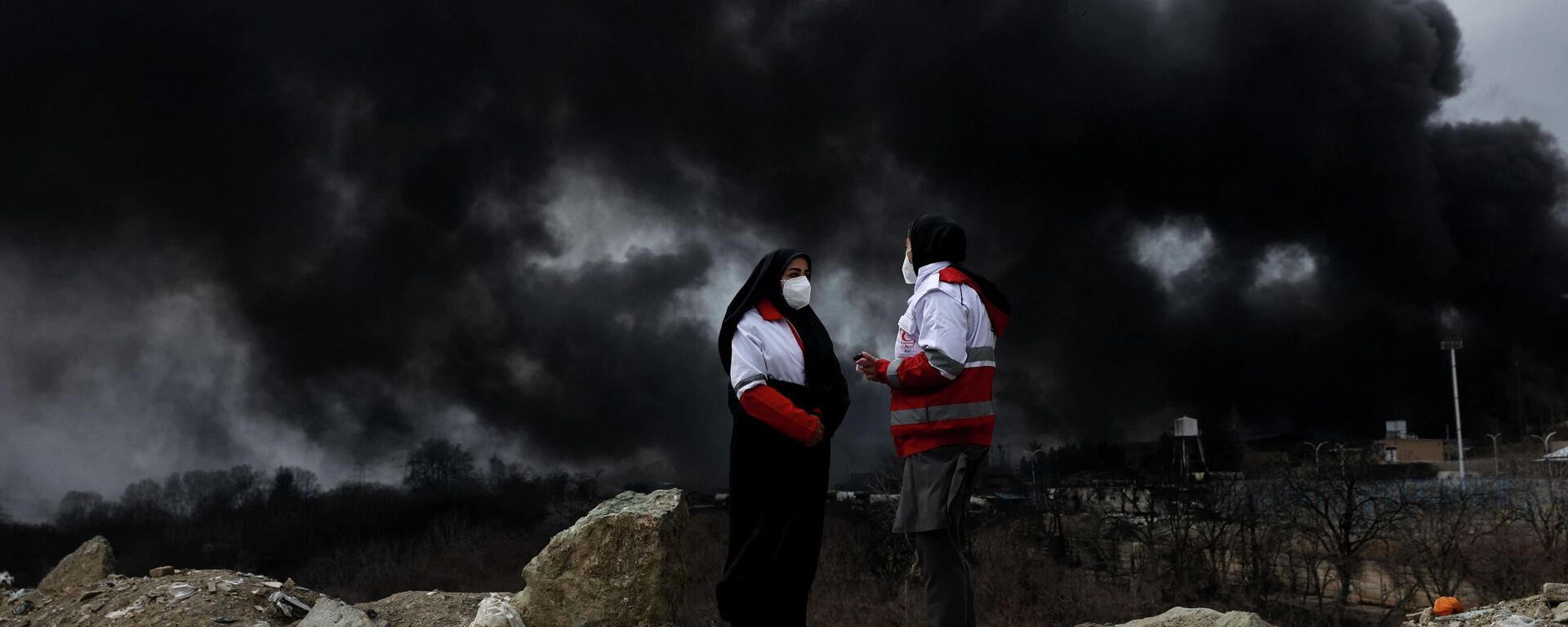 Các thành viên Hội Chữ thập đỏ nói chuyện với nhau khi khói bốc lên sau một vụ tấn công được báo cáo vào các bể chứa nhiên liệu ở Shahran, Iran - Sputnik Việt Nam, 1920, 12.03.2026